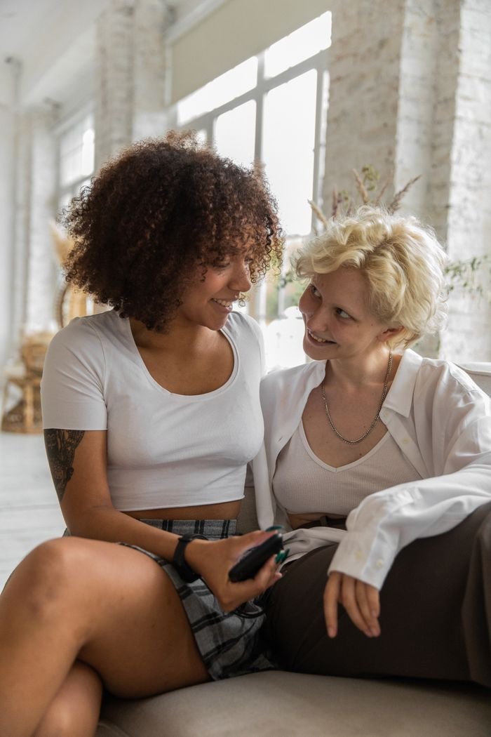 Young multiracial lesbian couple resting on sofa with mobile phone and embracing while looking at each other
