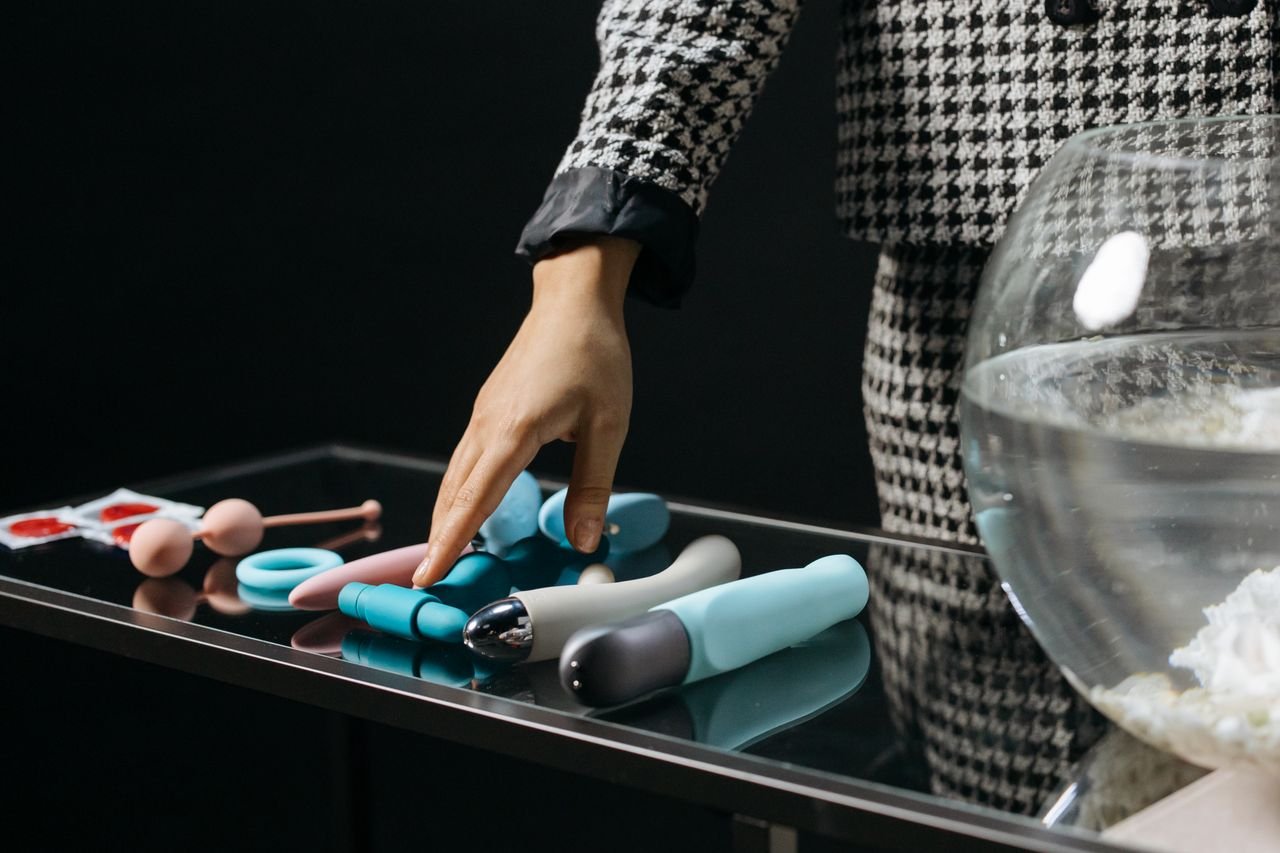 Hand reaching for various colorful silicone adult toys on a reflective glass table. Sensual and vibrant.